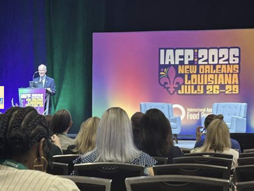 Speaker at a podium presenting on a brightly lit stage; IAFP 2026 banner behind him and seated audience in front.